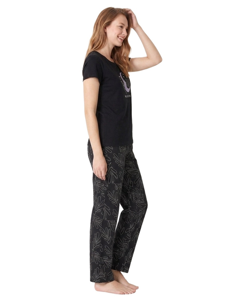 A woman modeling a two-piece pajama set consisting of a black short-sleeve t-shirt with a graphic print and grey pants featuring a light grey abstract leaf pattern, set against a plain white background.