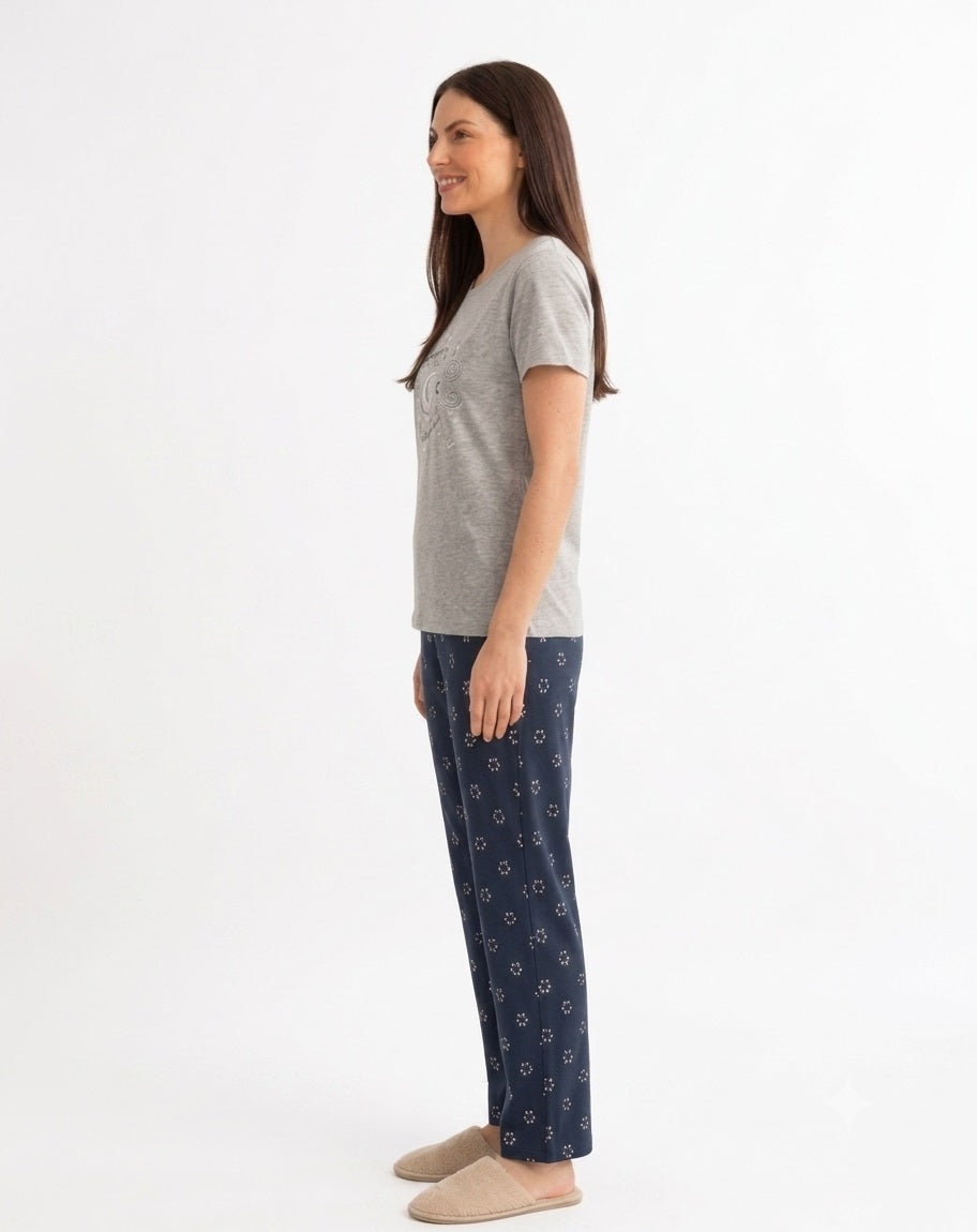A woman stands in profile wearing a gray melange short-sleeve round neck top and peacock blue pants with a small white floral pattern, paired with beige slippers, against a plain white studio background.