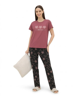 A woman modeling a two-piece pajama set consisting of a dusty pink short-sleeve t-shirt with a white floral graphic and black pants with a pink floral vine pattern, holding a white pillow against a plain white background.