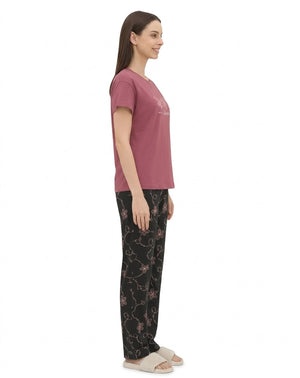 A woman shown in profile wearing a dusty pink short-sleeve pajama top and black pajama pants with a pink floral vine pattern, standing against a plain white background.